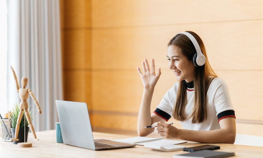 Smiling Asian young female using headset looking at laptop screen listen and learning online courses